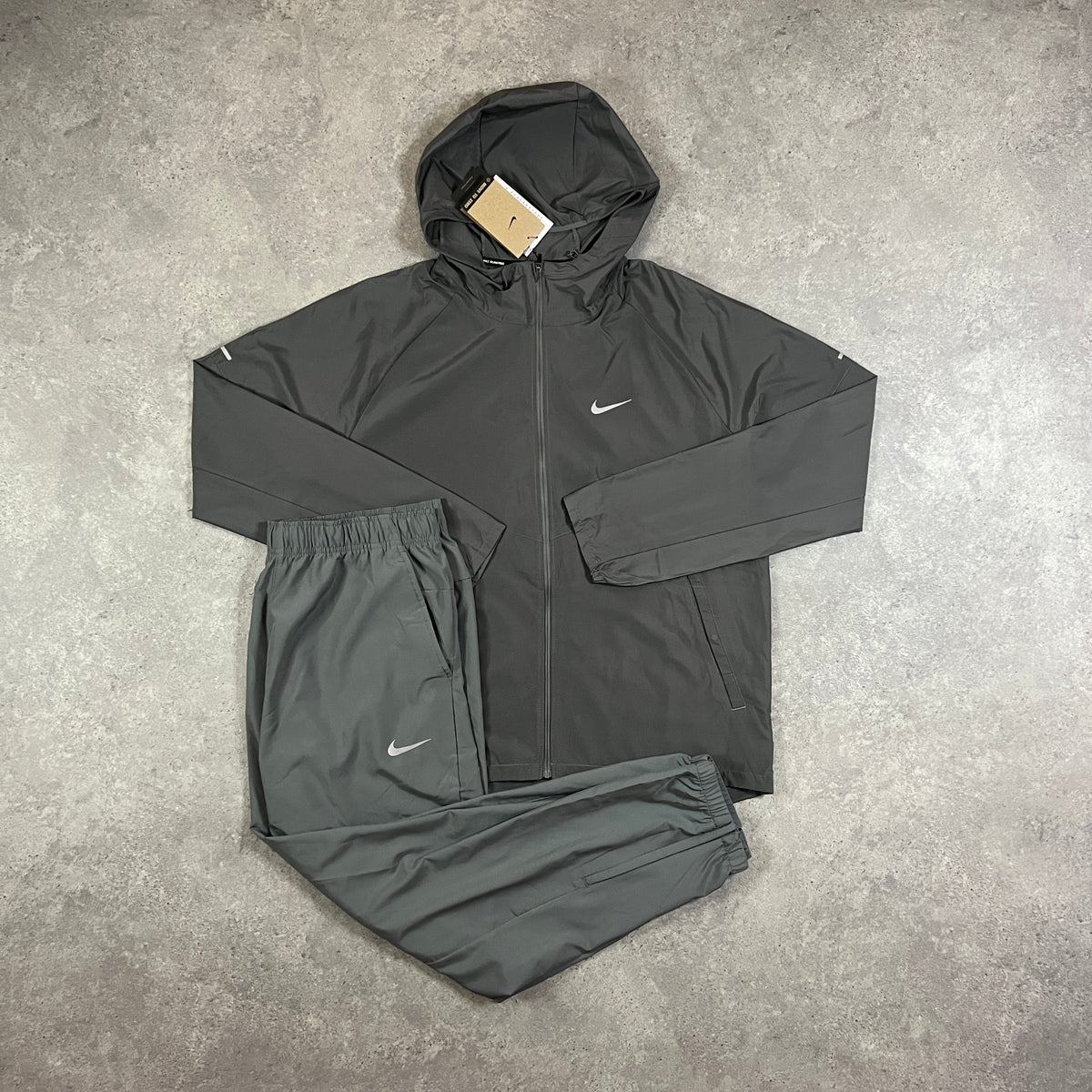 Nike Repel Tracksuit Grey – grey zip-up hooded windrunner jacket with white Nike Swoosh and reflective detailing, paired with matching grey joggers with Swoosh logo, flat lay on concrete surface with tags attached