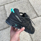 ASICS Gel Venture 6 Tiffany – all-black chunky trail-inspired trainer held in hand, heel view showing Gel Venture 6 branding, teal heel pull tab and teal GEL cushioning pod on black outsole against paving stones