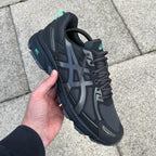 ASICS Gel Venture 6 Tiffany – all-black chunky trail-inspired trainer held in hand, top-down lateral view showing black mesh upper, dark ASICS Stripes, black laces and teal collar pull tab against paving stones