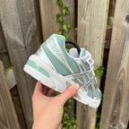 ASICS Gel 1130 Verdigris Pure Silver – sage green and silver retro trainer held in hand, heel view showing Gel 1130 branding and chunky white outsole with mint GEL pod against wooden fence