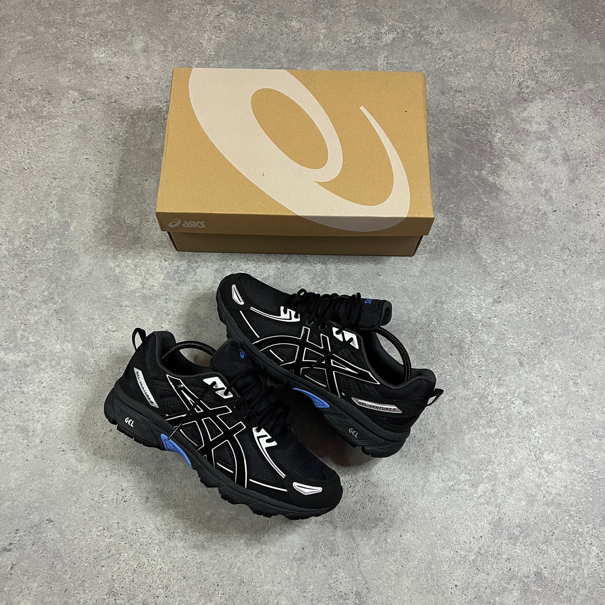 ASICS Gel Venture 6 Blue Coast – pair of black chunky trail trainers with white ASICS Stripes and blue GEL accents, flat lay top-down view alongside ASICS branded shoebox on concrete surface