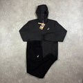 ASICS Core Windrunner & Core Bottoms Black – black zip-up hooded windrunner jacket with white ASICS chest logo, paired with matching black core bottoms with ASICS logo, flat lay on concrete surface with tags attached
