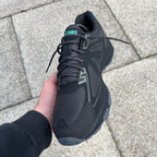 ASICS Gel Venture 6 Tiffany – all-black chunky trail-inspired trainer held in hand, front top-down view showing black mesh toe box, black laces and teal ASICS logo on tongue against paving stones